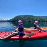 Two people paddling red and orange kayaks on a lake with a snow-capped mountain backdrop.