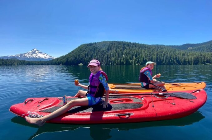 Two people paddling red and orange kayaks on a lake with a snow-capped mountain backdrop.