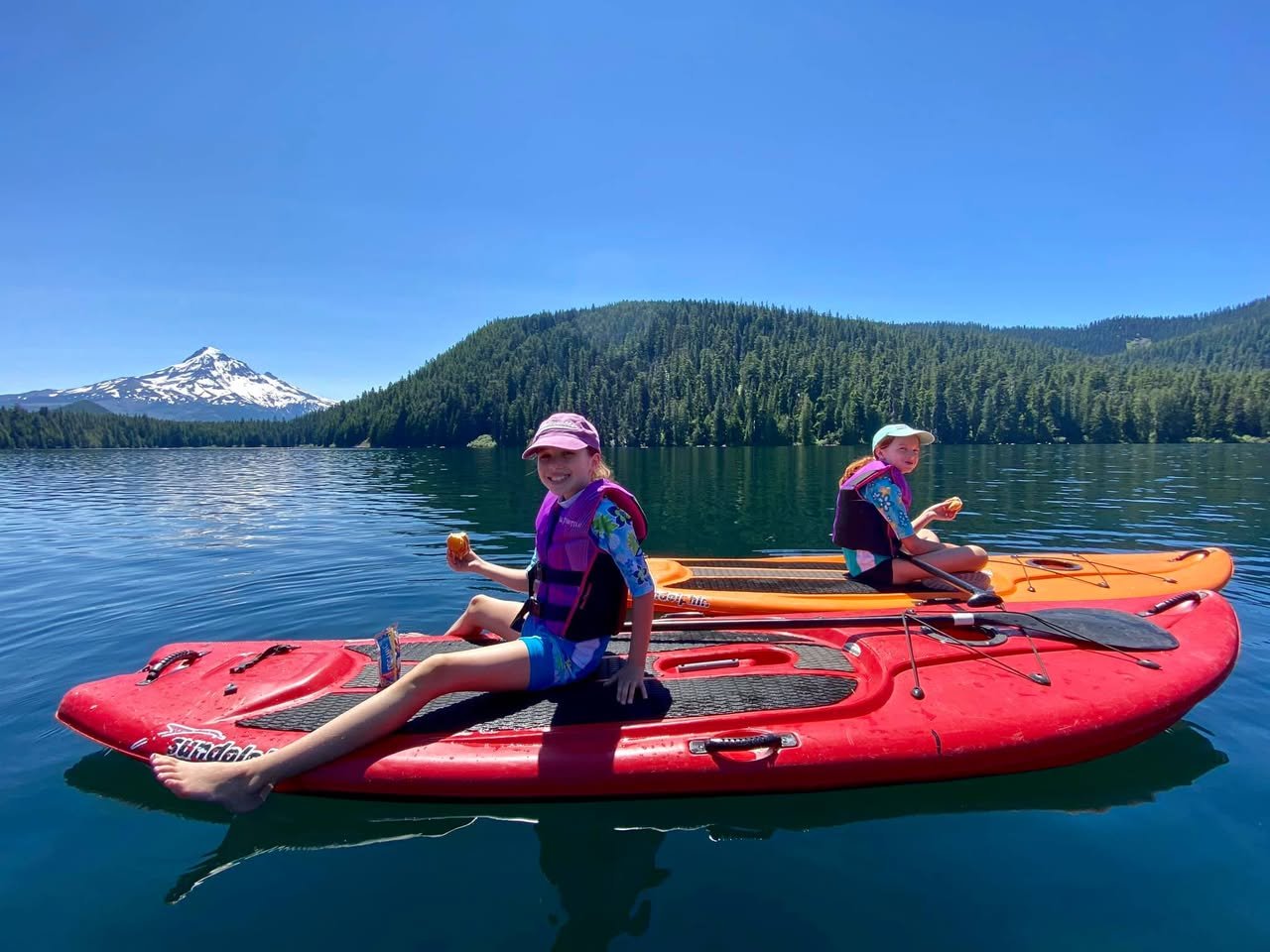 Two people paddling red and orange kayaks on a lake with a snow-capped mountain backdrop.