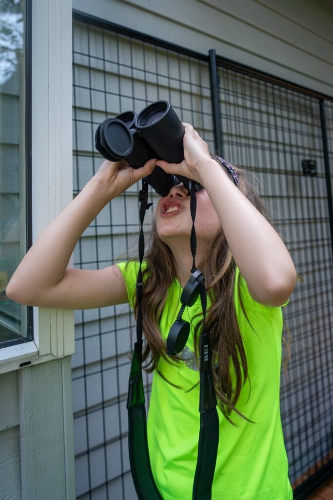 A person in a bright green shirt holding black binoculars to their eyes while looking upward outdoors