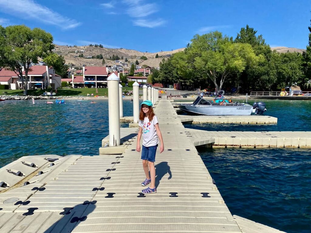 A scenic view of a wooden dock extending into a clear blue lake with a moored boat, surrounded by lush green trees and a residential shoreline. A person walks along the dock under a bright sunny sky.