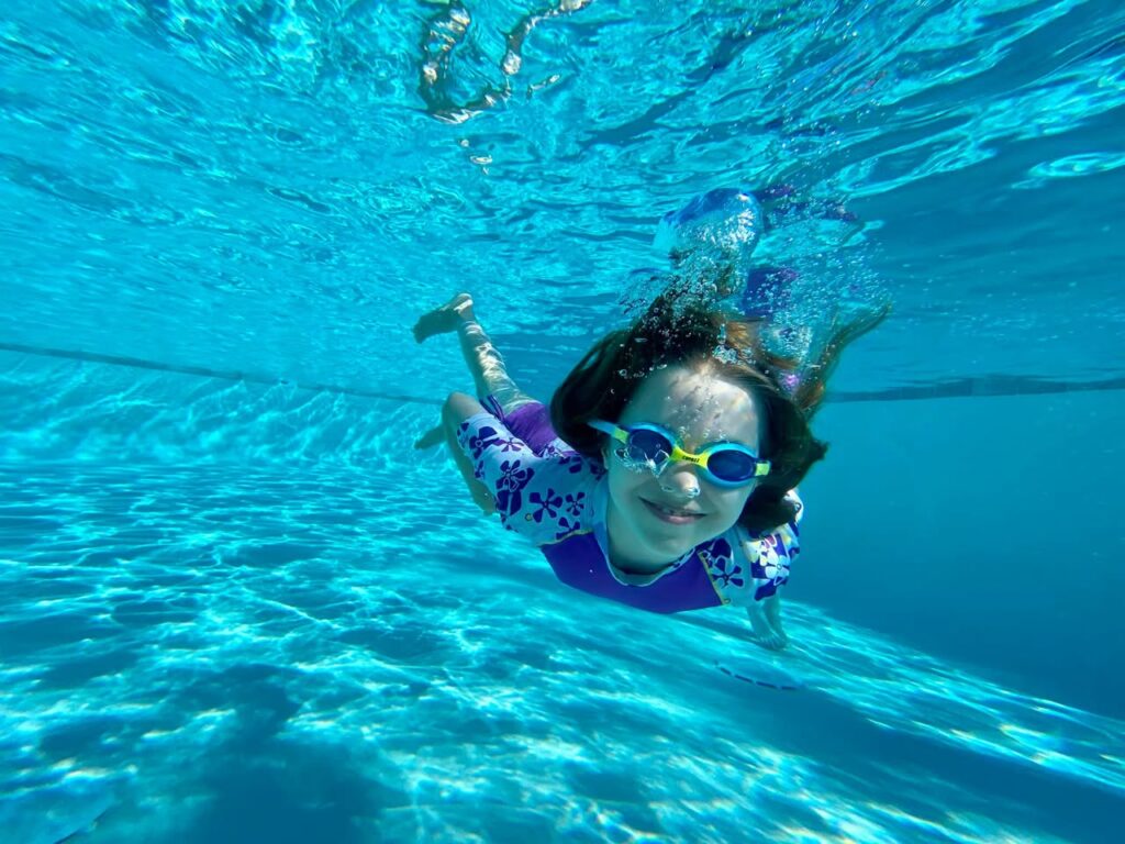 A person wearing a life vest and goggles swims underwater in a bright blue pool. Sunlight patterns dance across the water surface above.