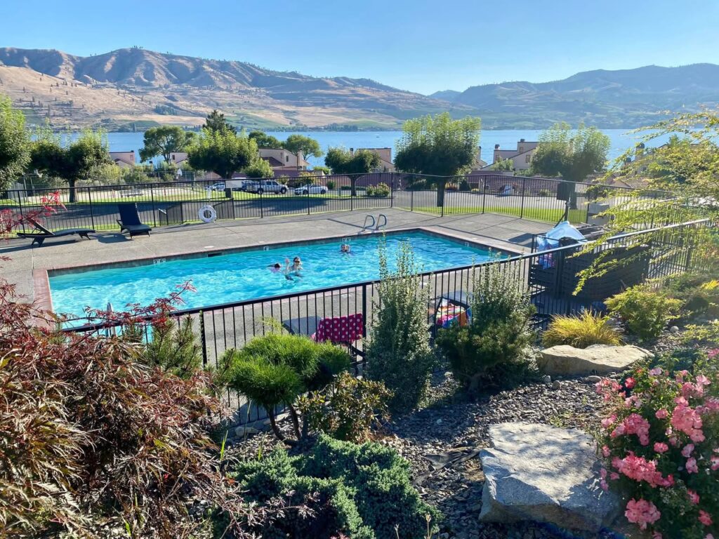 An elevated view of a motel pool with people swimming, overlooking a lake, trees, and distant arid mountains under a clear blue sky.