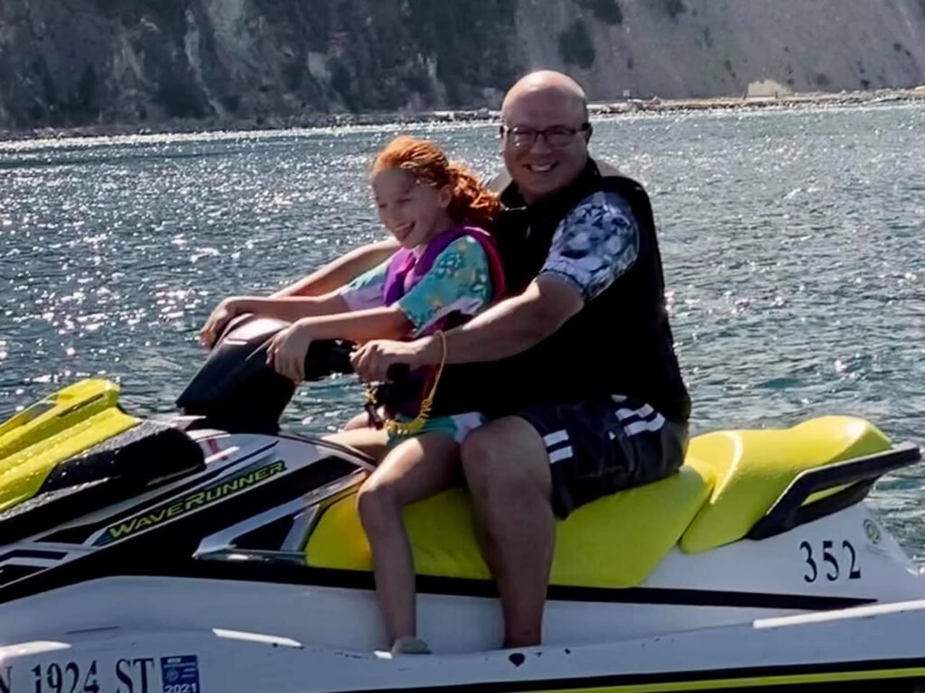 A smiling adult and child ride a yellow and black jetski on sparkling blue water near rocky shoreline.