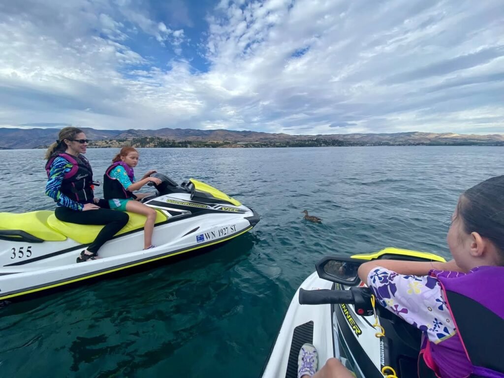 People ride jet skis on a vast lake surrounded by mountains under a partly cloudy sky.