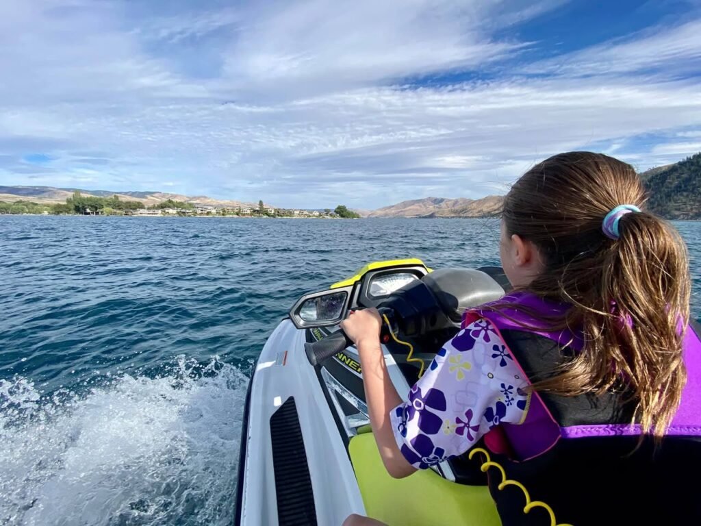 View from a jet ski showing a rider wearing a life vest splashing through blue water with a distant shoreline under a partly cloudy sky.