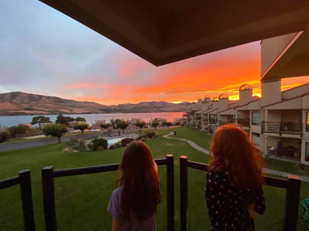 Two people watch a vibrant orange sunset over a lake from a multi-story resort balcony. The background features green lawns and distant hills under a colorful sky.