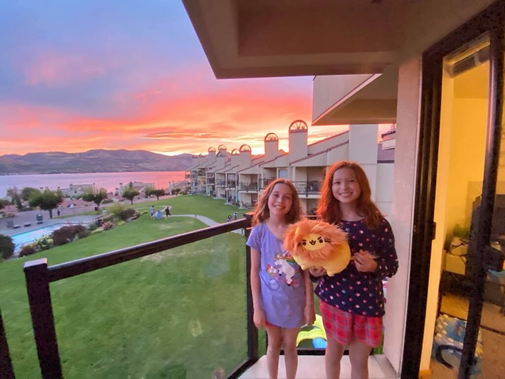 Two people holding a stuffed animal on a balcony with a scenic lake and sunset view at dusk.