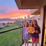 Two people holding a stuffed animal on a balcony with a scenic lake and sunset view at dusk.