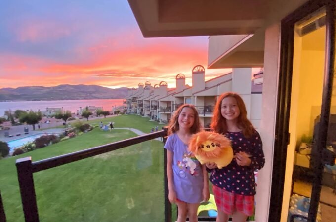 Two people holding a stuffed animal on a balcony with a scenic lake and sunset view at dusk.