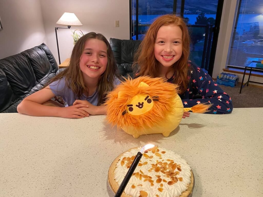A cozy indoor birthday celebration featuring two smiling young friends, a lion plushie, and a dessert with a lit candle.
