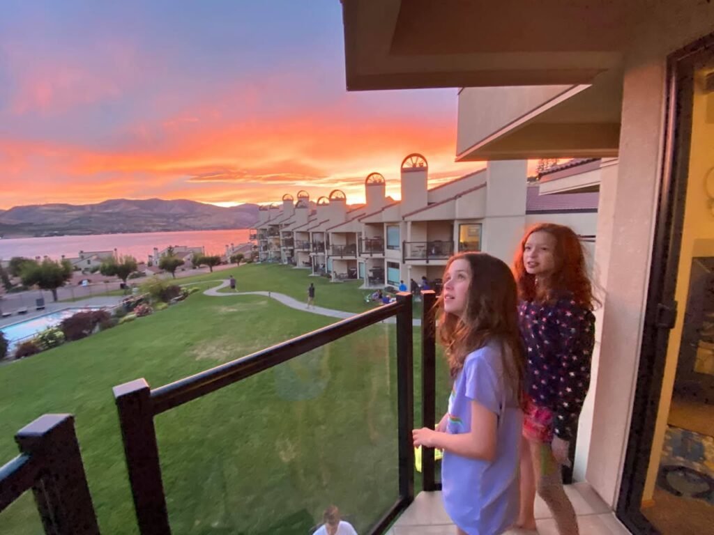 Two individuals on a balcony enjoy the view of a golf course and a lake during a vibrant sunset.