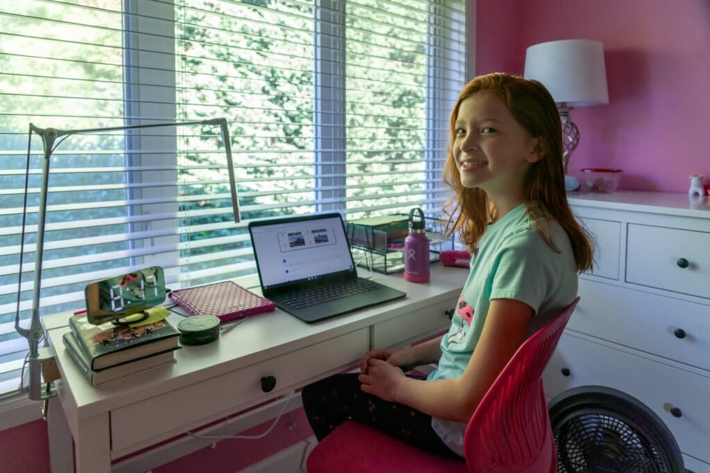 A young person using a laptop at a white desk with a view of blinds and trees.