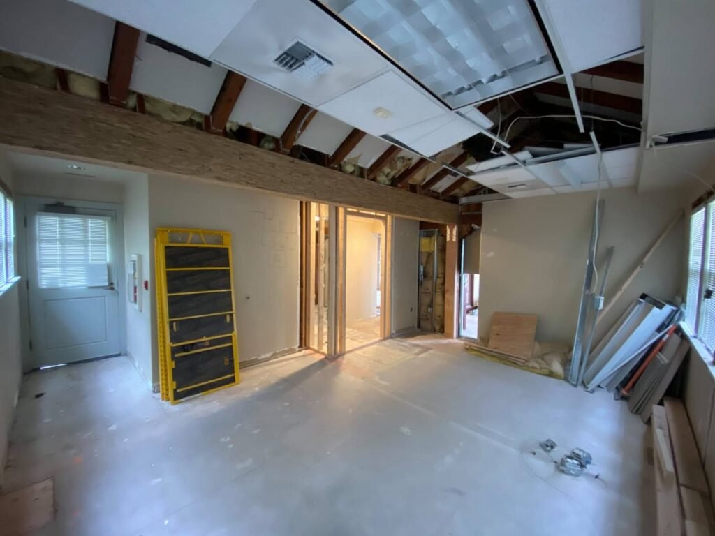A room under renovation with exposed ceiling beams and unfinished walls. Construction materials are visible, including a yellow shelving unit and a ladder.