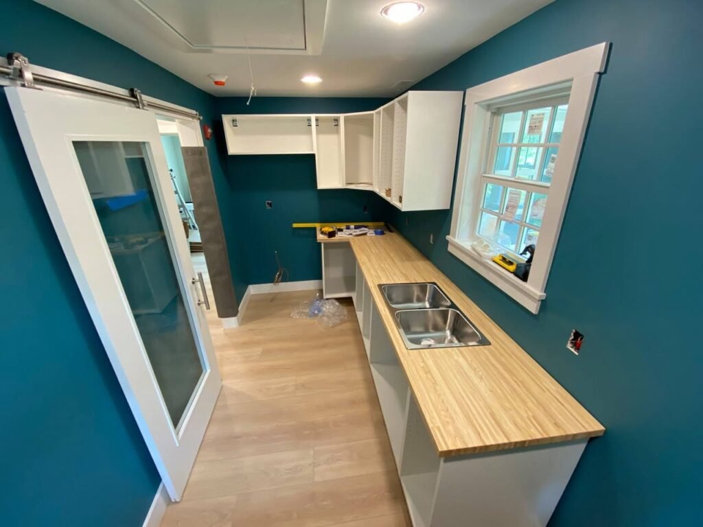 Bright kitchen with teal walls, white cabinets, and a light wood countertop featuring a stainless steel sink. A sliding glass door is visible beside the long counter.