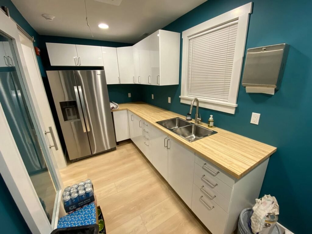 A kitchen with dark teal walls, white cabinets, and light wood countertops, featuring a stainless steel double sink and large refrigerator.