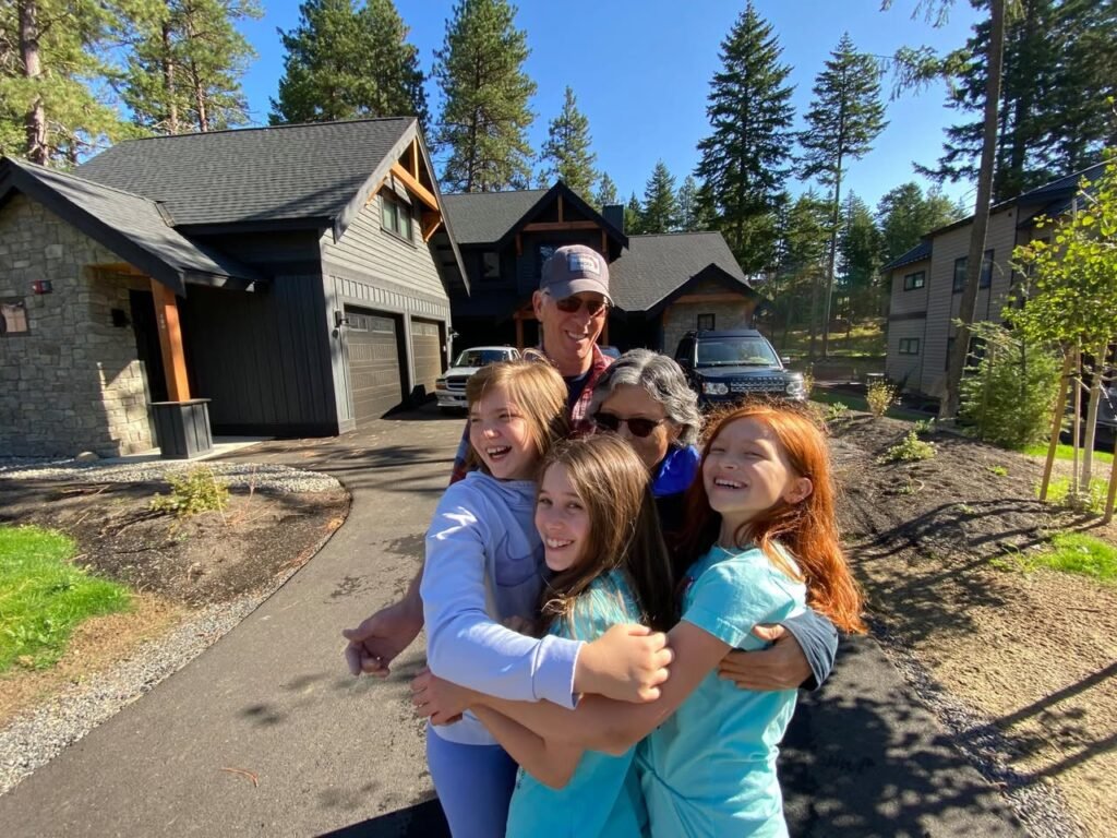 A family group hugs in front of a mountain cabin surrounded by tall evergreen trees.
