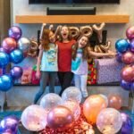 Three girls celebrating a birthday with metallic balloons and a 'Happy Birthday' sign.