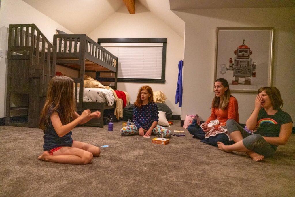 A group of people sits on the floor in a living room playing cards and board games next to a bunk bed.