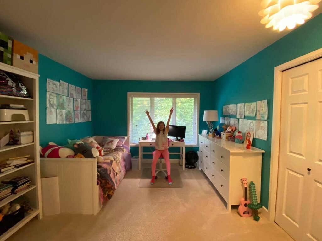 A child playfully jumping in a teal-walled bedroom with white furniture. The room features a desk, and clothing poster.