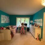 A child playfully jumping in a teal-walled bedroom with white furniture. The room features a desk, and clothing poster.