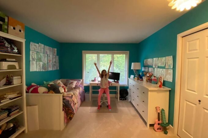 A child playfully jumping in a teal-walled bedroom with white furniture. The room features a desk, and clothing poster.