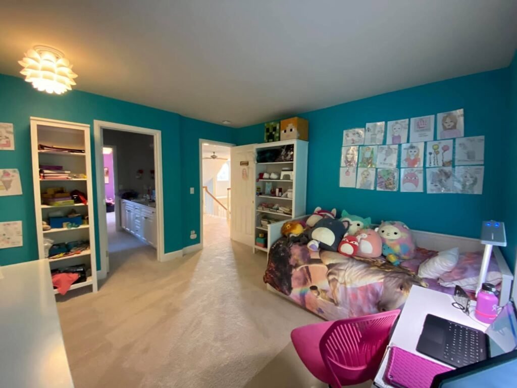 A bright bedroom featuring teal walls, built-in shelves, and a bed with plush toys. A laptop on a white desk adds a modern touch to the cozy space.