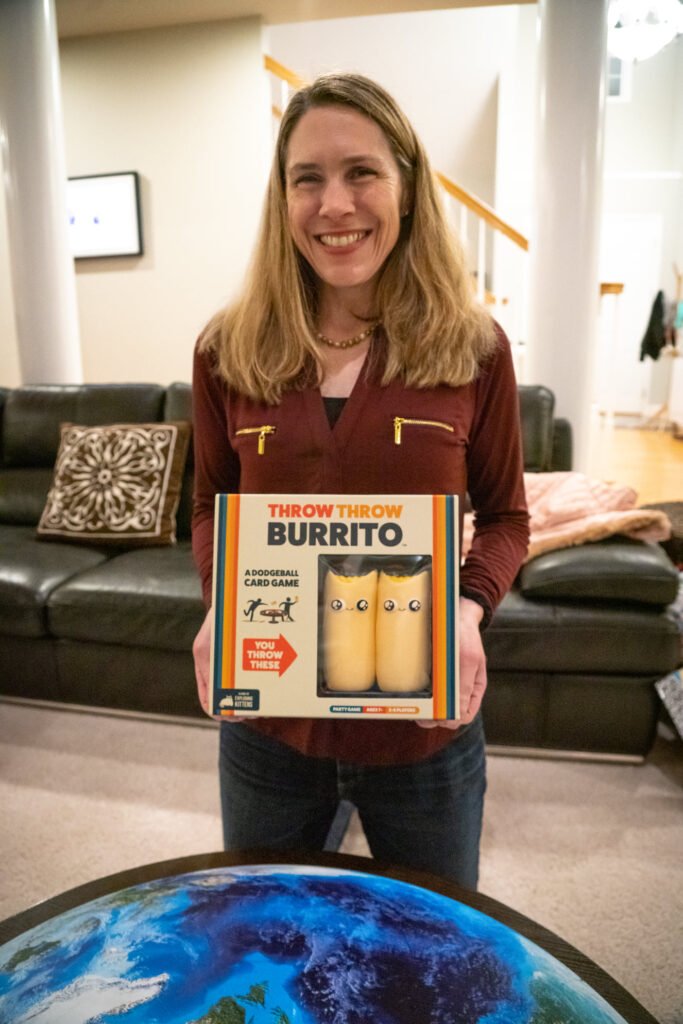 A person stands in a living room holding a box labeled Burrito, with a globe-coffee table and a dark couch behind.