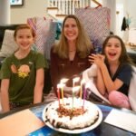 Three people smiling around a birthday cake with lit candles on a coffee table. Cozy living room scene with wrapped gifts and a black leather sofa.