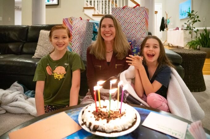 Three people smiling around a birthday cake with lit candles on a coffee table. Cozy living room scene with wrapped gifts and a black leather sofa.