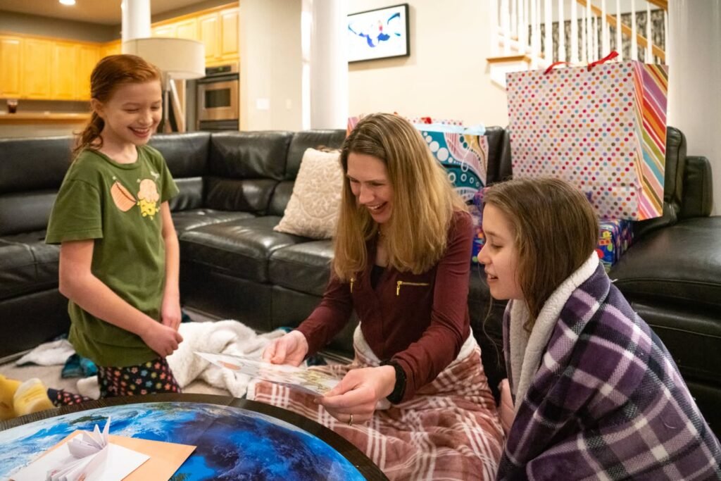 A grandmother and two grandchildren opening gifts together in a warm, festive home setting. A colorful globe table adds a playful touch to the family tradition.