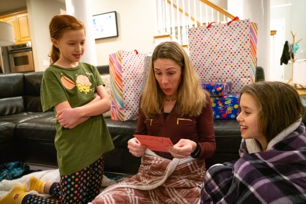 A close-up of a person sitting on a black leather couch, wrapped in a plaid blanket, surrounded by wrapped presents.