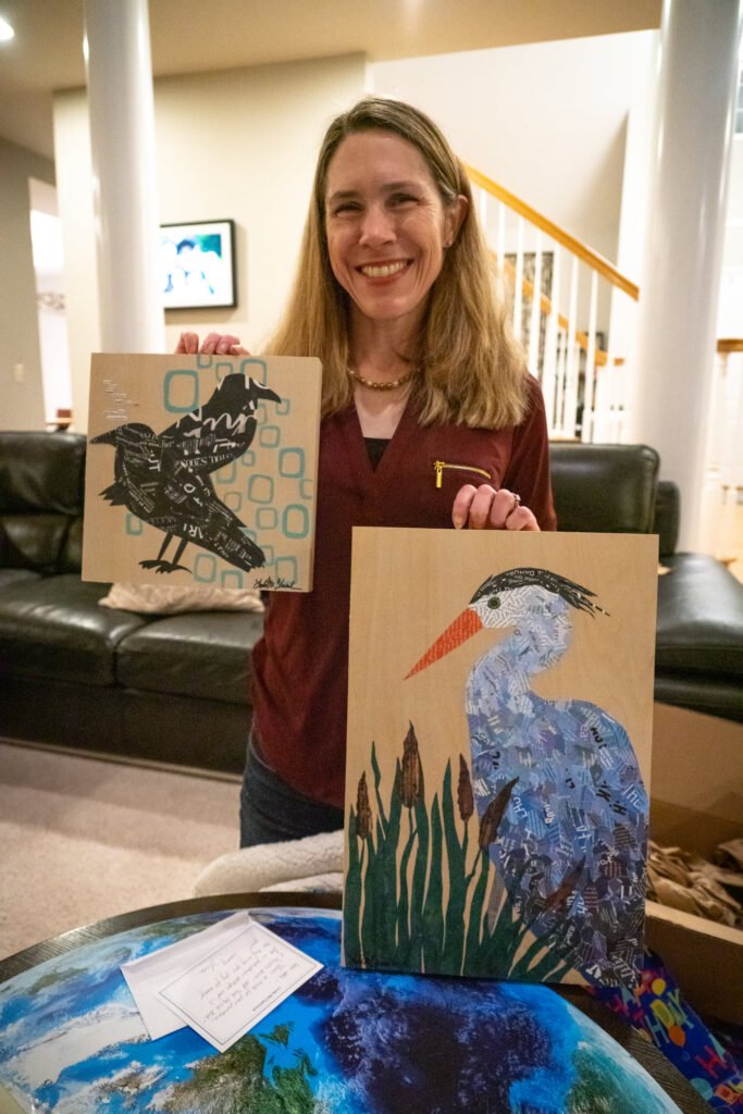 A woman smiles while holding two canvas paintings featuring a cormorant and two black birds.
