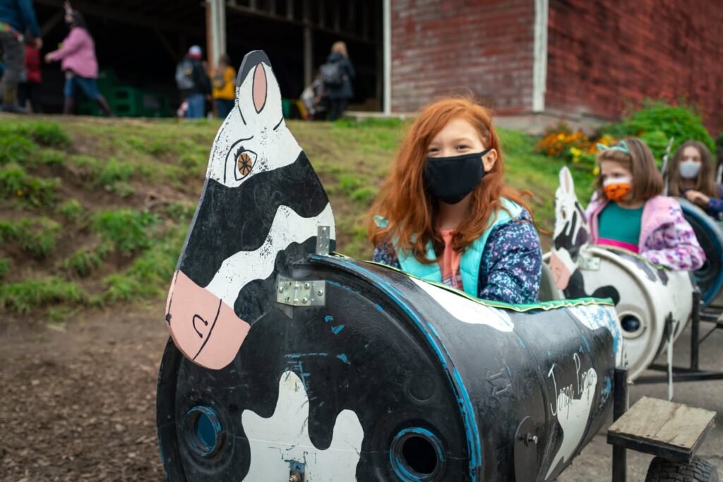 A group of people wearing masks rides a cow-shaped carousel ride outdoors. The setting appears to be a park or zoo with brick buildings and grass nearby.