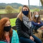 19th Anniversary Three individuals pictured seated outdoors in a rural landscape, wearing protective face masks. The backdrop features green fields and a farm environment with wooden fencing.