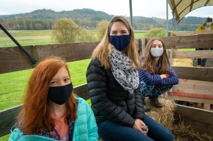 Three individuals pictured seated outdoors in a rural landscape, wearing protective face masks. The backdrop features green fields and a farm environment with wooden fencing.