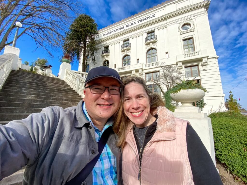 A selfie taken by a couple in front of a large white building with classical architecture under a blue sky.