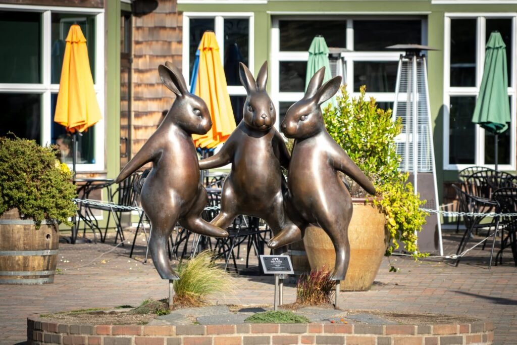 A trio of bronze rabbit sculptures dance on a brick patio in a sunny outdoor seating area beside a building with green walls. Colorful closed umbrellas and dining furniture add charm to the scene.