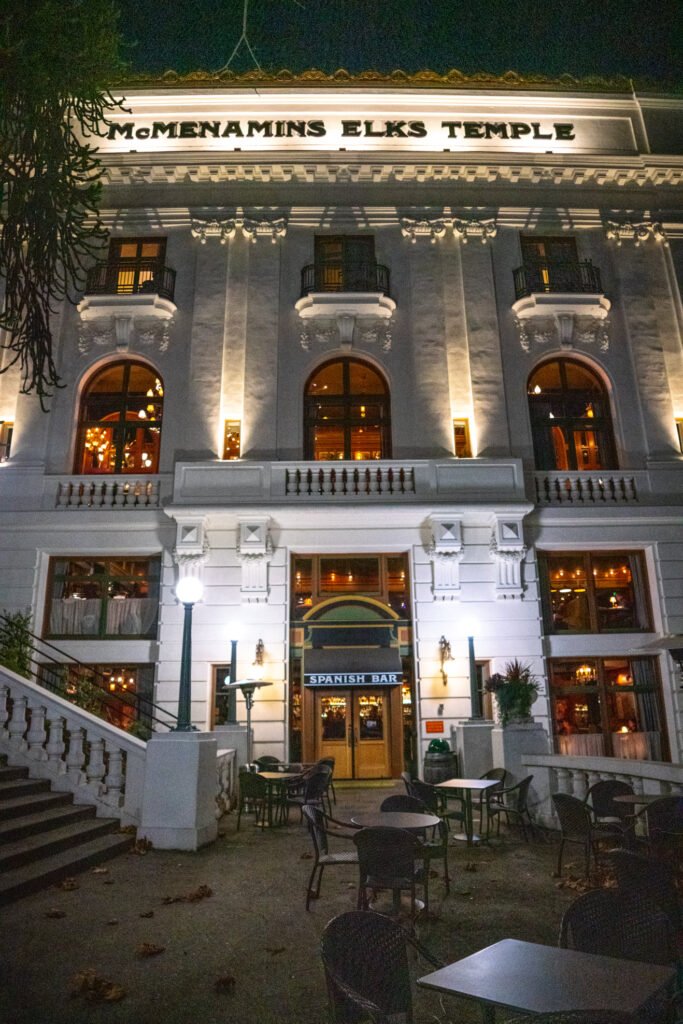 Nighttime view of the McMenamins Elks Temple building with illuminated windows and entrance. The exterior features classic architecture and outdoor seating area.