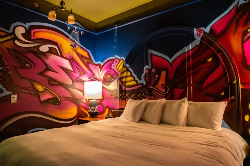 A bed with cream linens framed by vivid, abstract wall murals and unique pendant lighting.