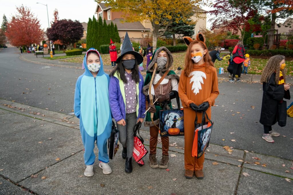 Children in Halloween costumes and masks trick-or-treating on a suburban sidewalk with autumn trees in the background.