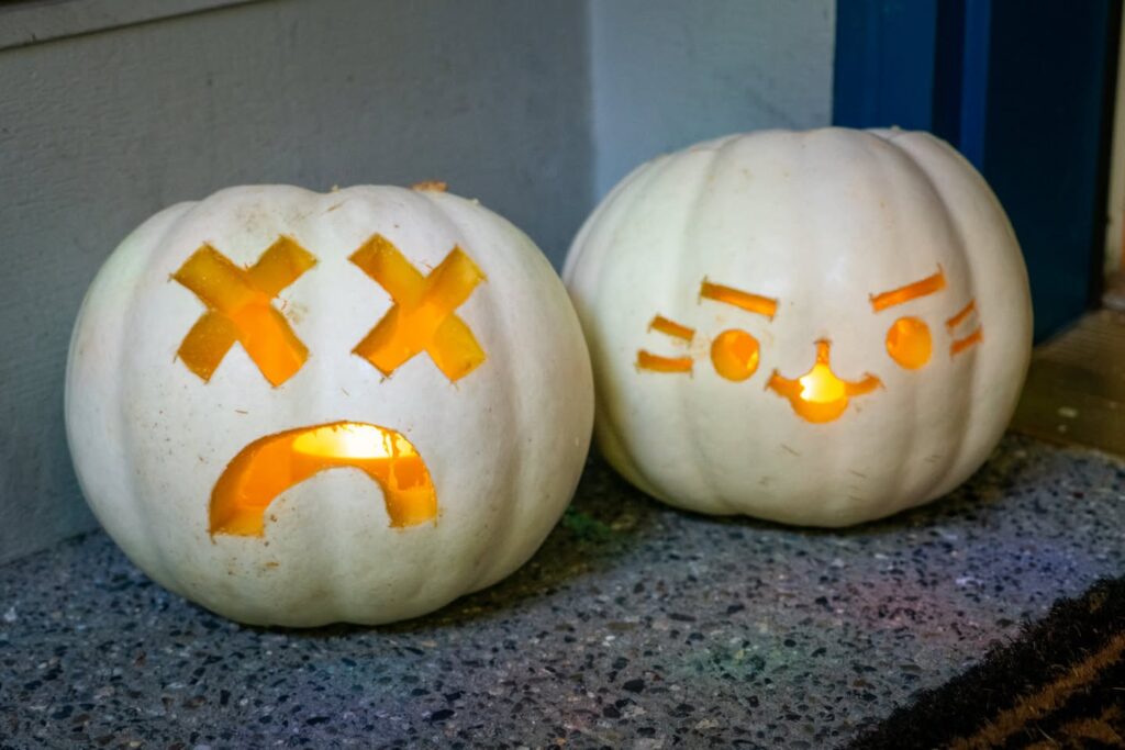 A white Halloween pumpkin carved with an X-eyed zombie face sits next to a smaller carved pumpkin with a friendly face.