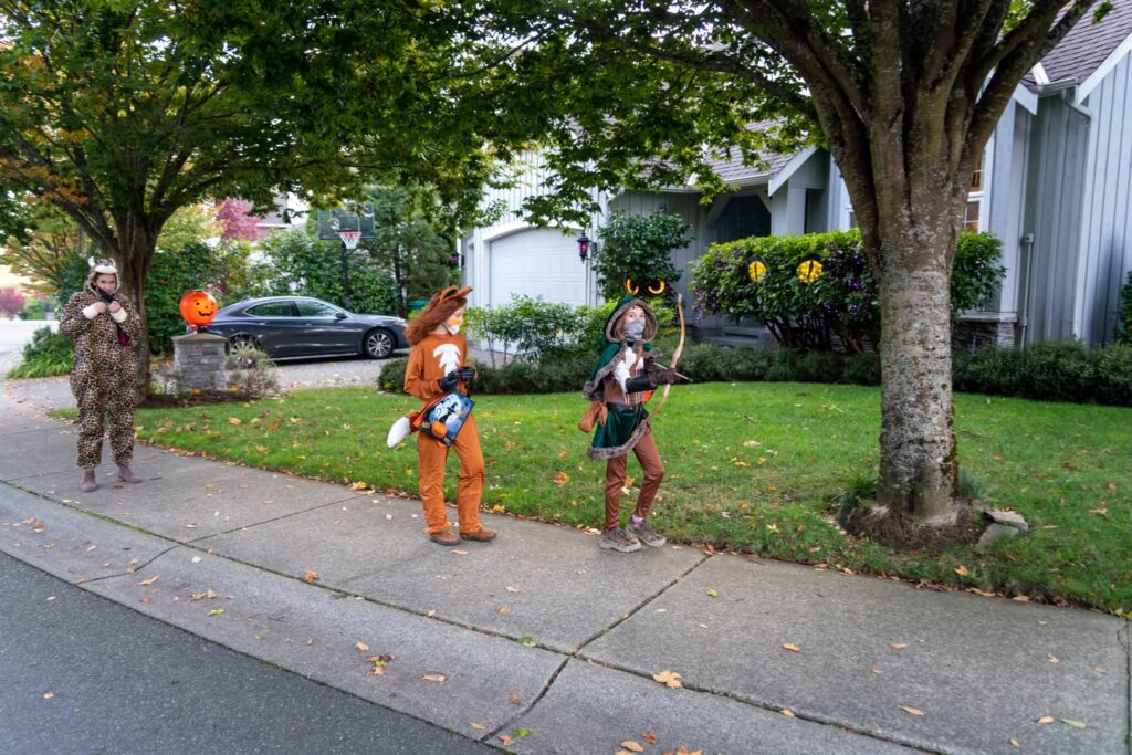 Individuals walking in costumes on a residential sidewalk lined with green lawns and mature trees.