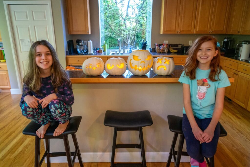 Two girls sitting on kitchen stools with carved and lit pumpkins on the counter behind them.