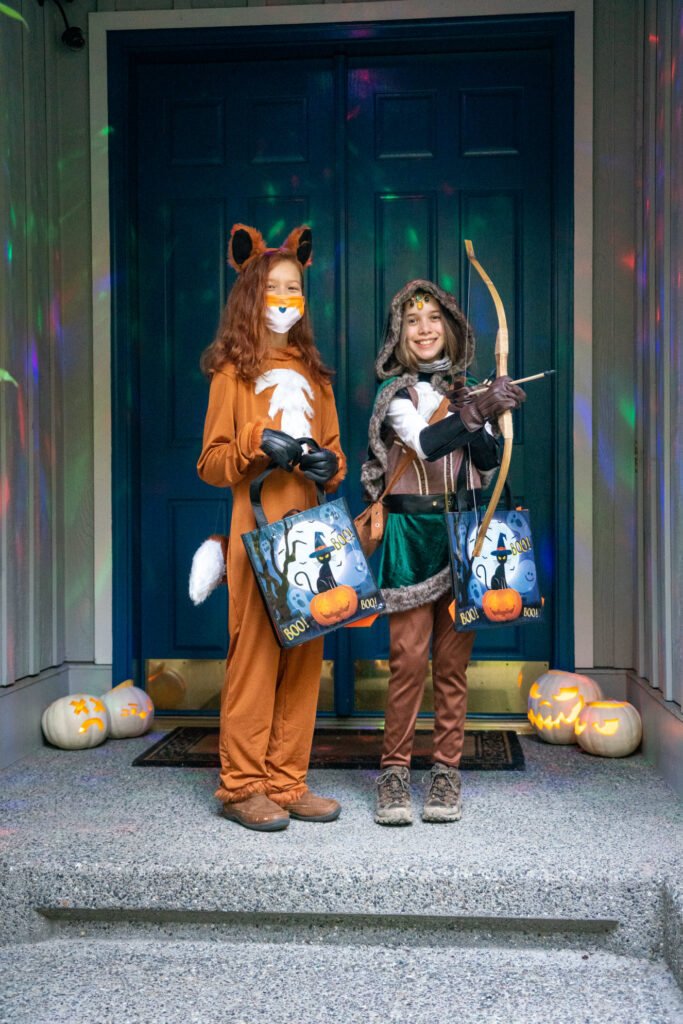 Two individuals in a Squirrel costume and an Archer costume stand on a porch step holding trick-or-treat bags.