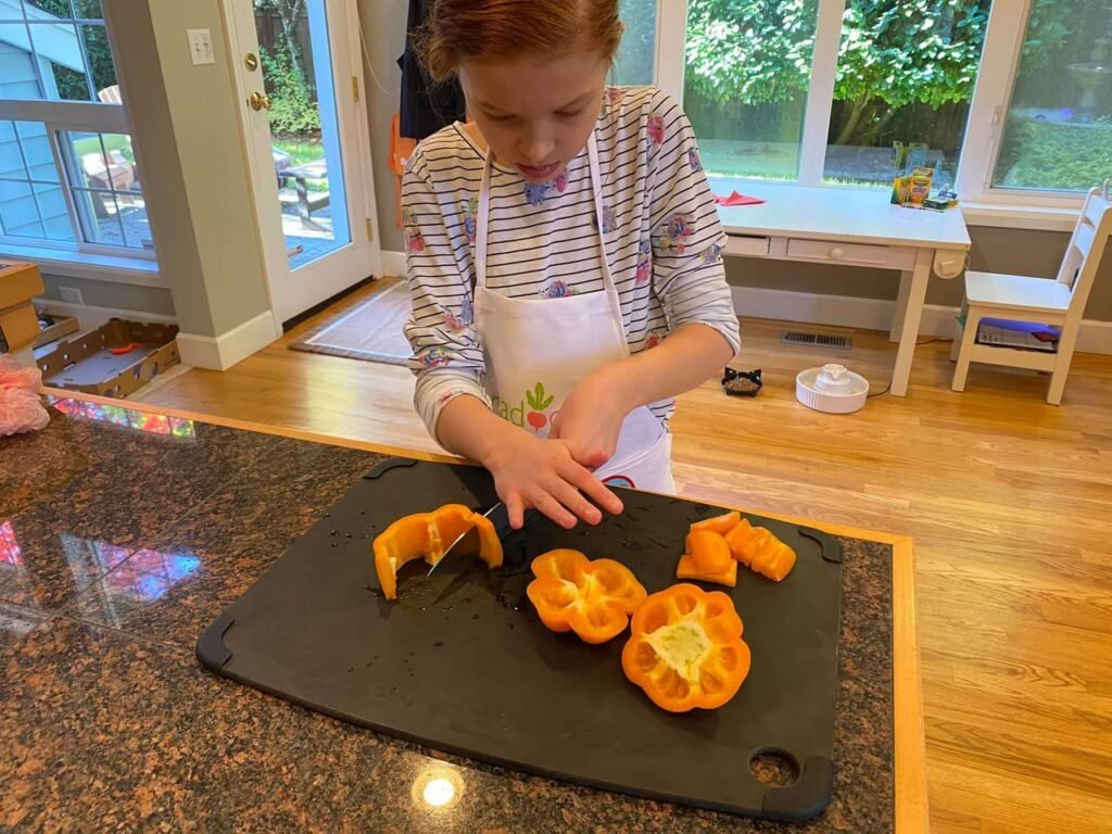 A child wearing an apron carefully slices yellow-orange fruit on a black cutting board. The background shows a bright kitchen with a view of the outdoors.