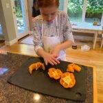 Bye Disco, Hello Mando A child wearing an apron carefully slices yellow-orange fruit on a black cutting board. The background shows a bright kitchen with a view of the outdoors.
