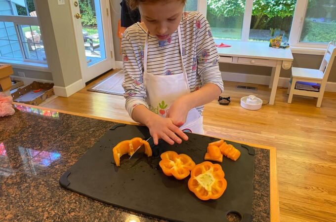 A child wearing an apron carefully slices yellow-orange fruit on a black cutting board. The background shows a bright kitchen with a view of the outdoors.