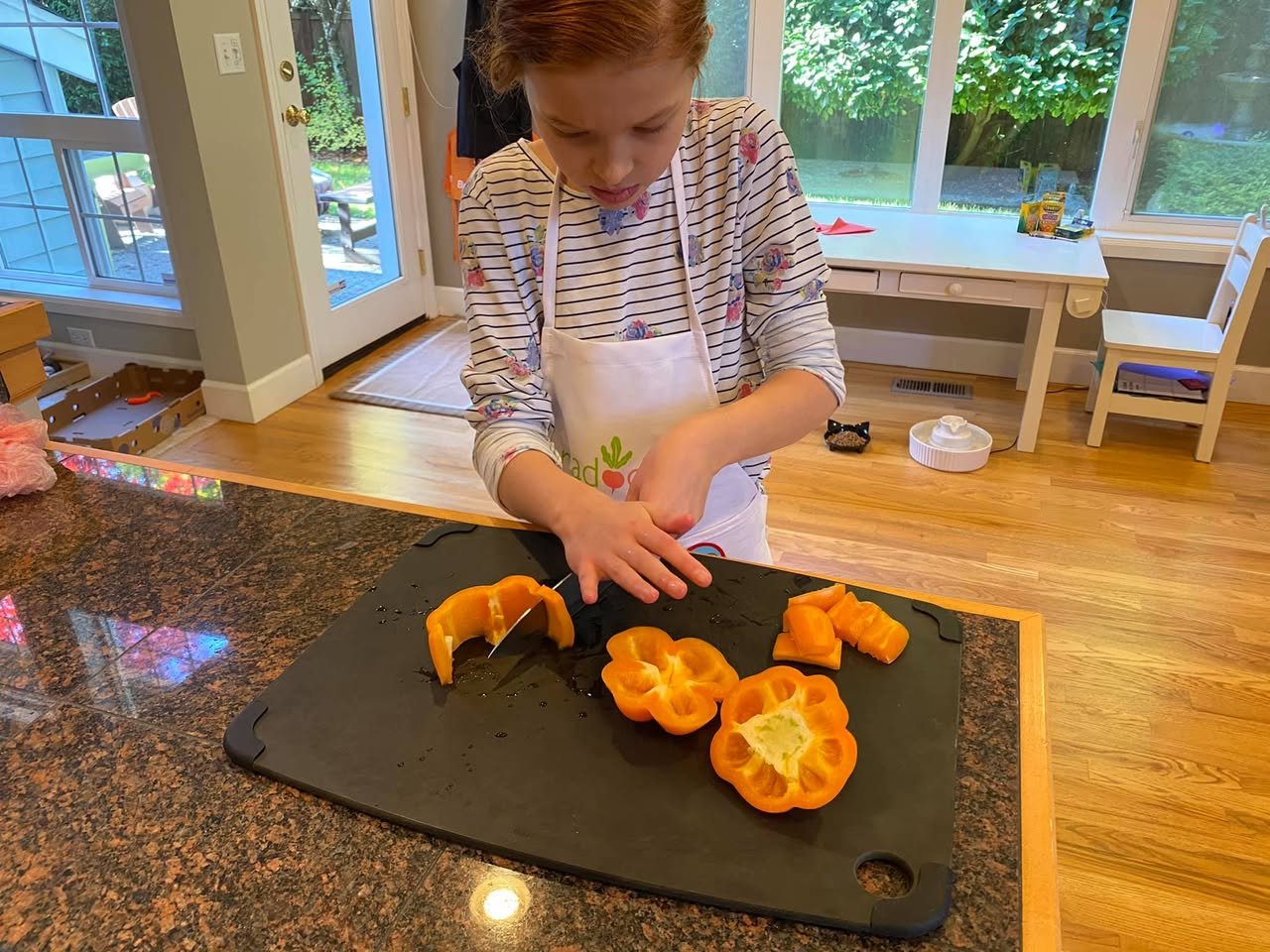 A child wearing an apron carefully slices yellow-orange fruit on a black cutting board. The background shows a bright kitchen with a view of the outdoors.