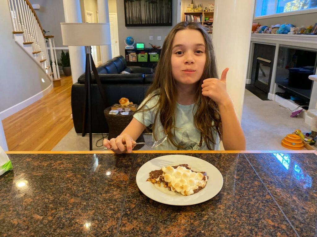 A young girl gives a thumbs-up while seated at a granite kitchen island. A plate of food is in front of her in a bright, open-plan living area with stairs and a fireplace visible.
